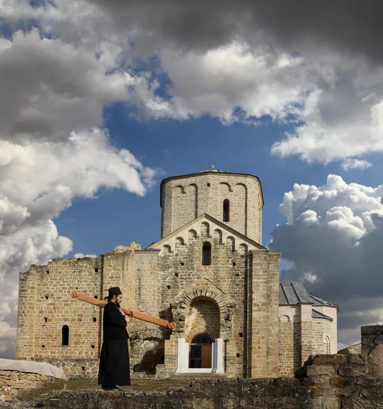 Srednjovekovni manastir sa dva kamena zvonika na zelenom brežuljku, plavo nebo.
